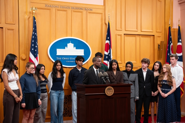 Senator Willis Blackshear Jr speaking at a podium with a group of student advocates standing behind him.